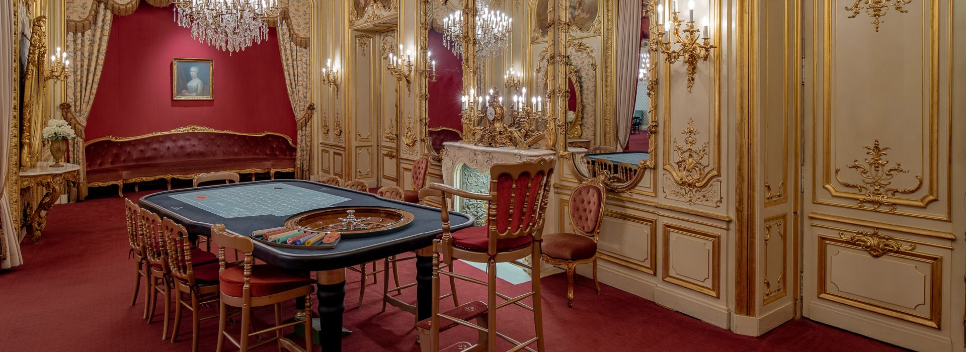 Luxurious casino room with chandeliers, gaming table, mirrors, and rich red decor