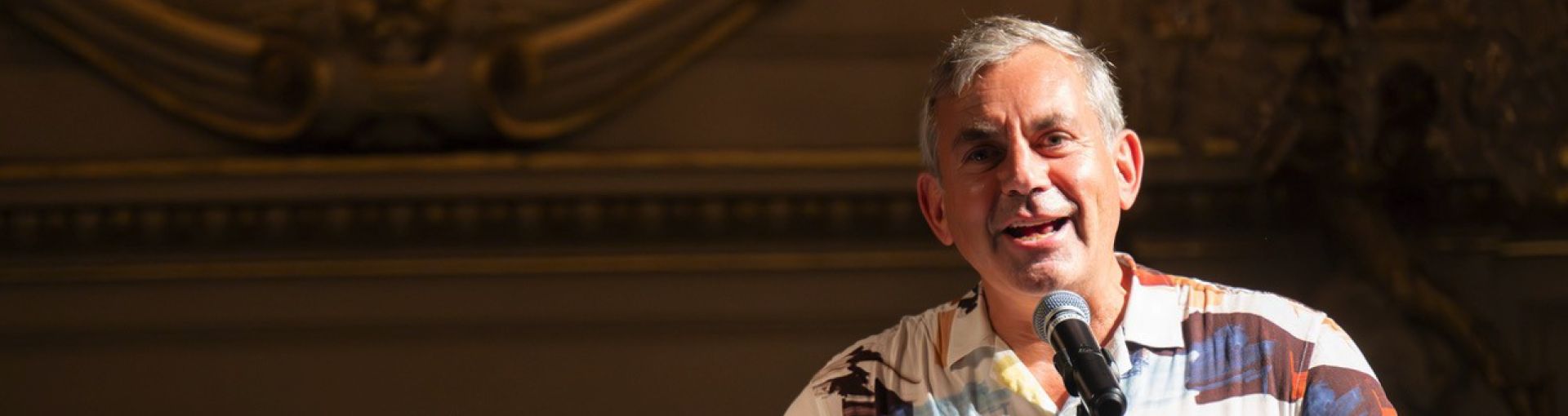 Person speaking into microphone in colorful shirt at event with decorative backdrop