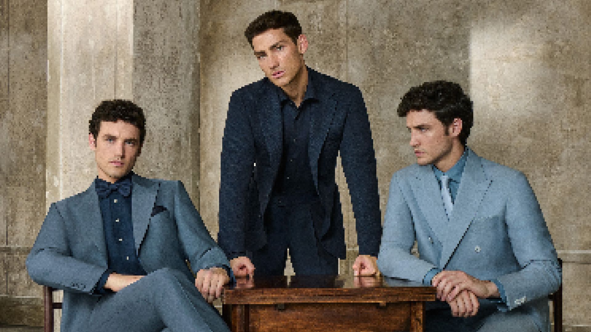 Three men in tailored suits posing around a wooden table indoors