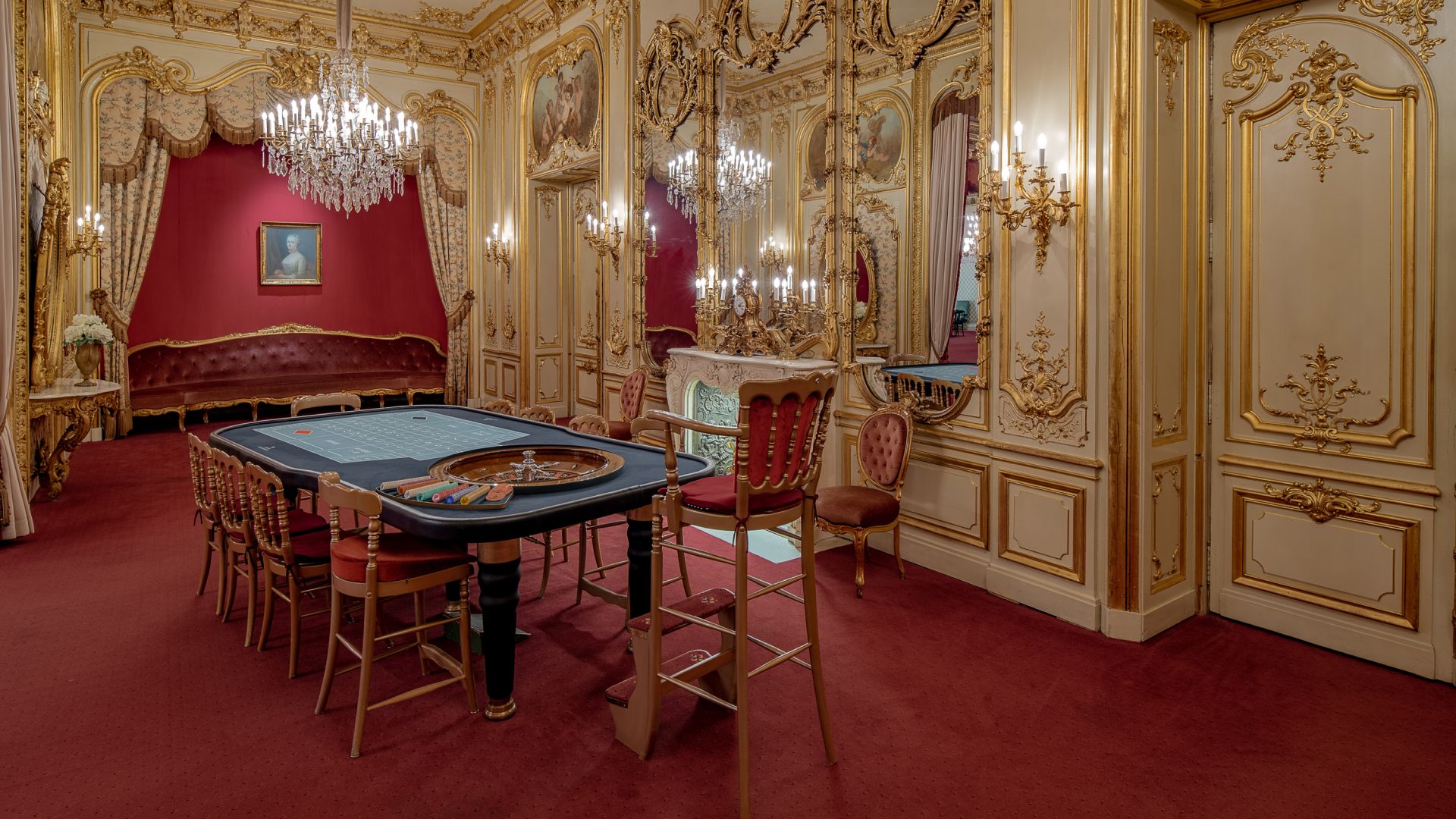 Luxurious room with a gambling table, gold decor, chandeliers, and plush carpeting.